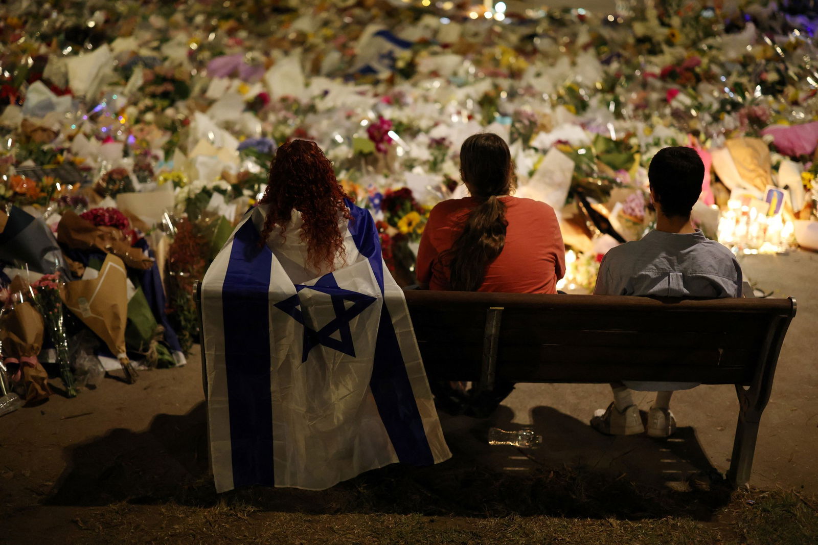 Três pessoas sentam-se num banco em frente a uma grande pilha de flores e uma mulher carrega uma bandeira israelense.