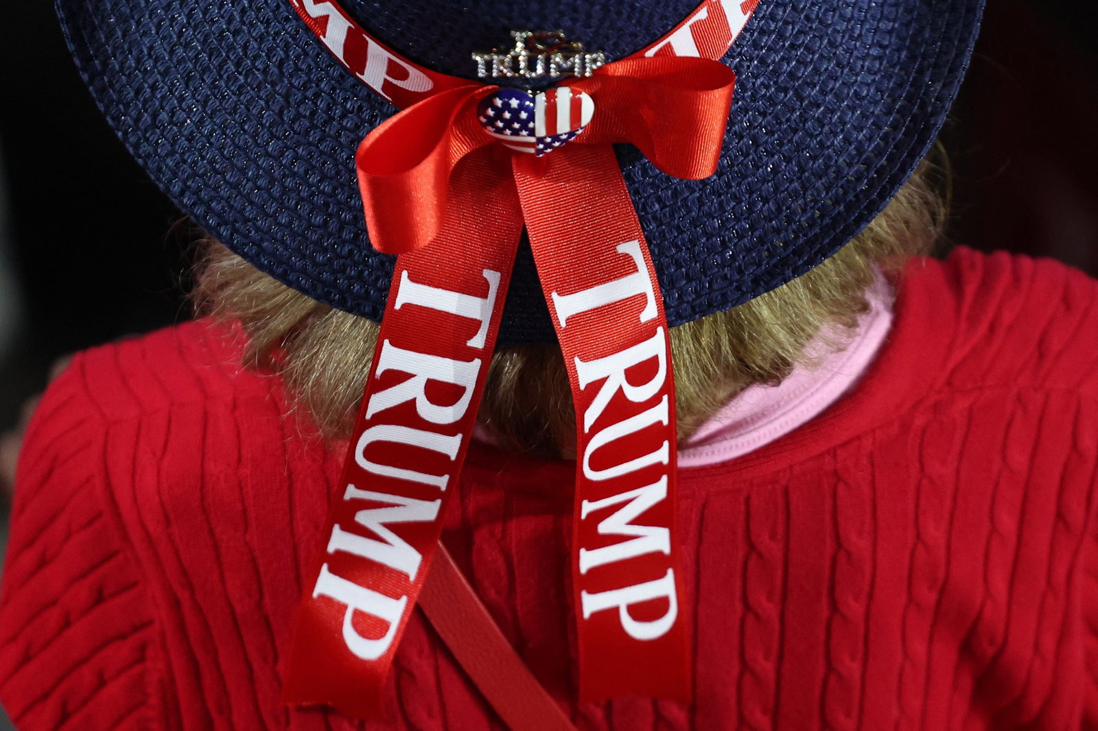 A woman wears a hat with a red ribbon saying Trump