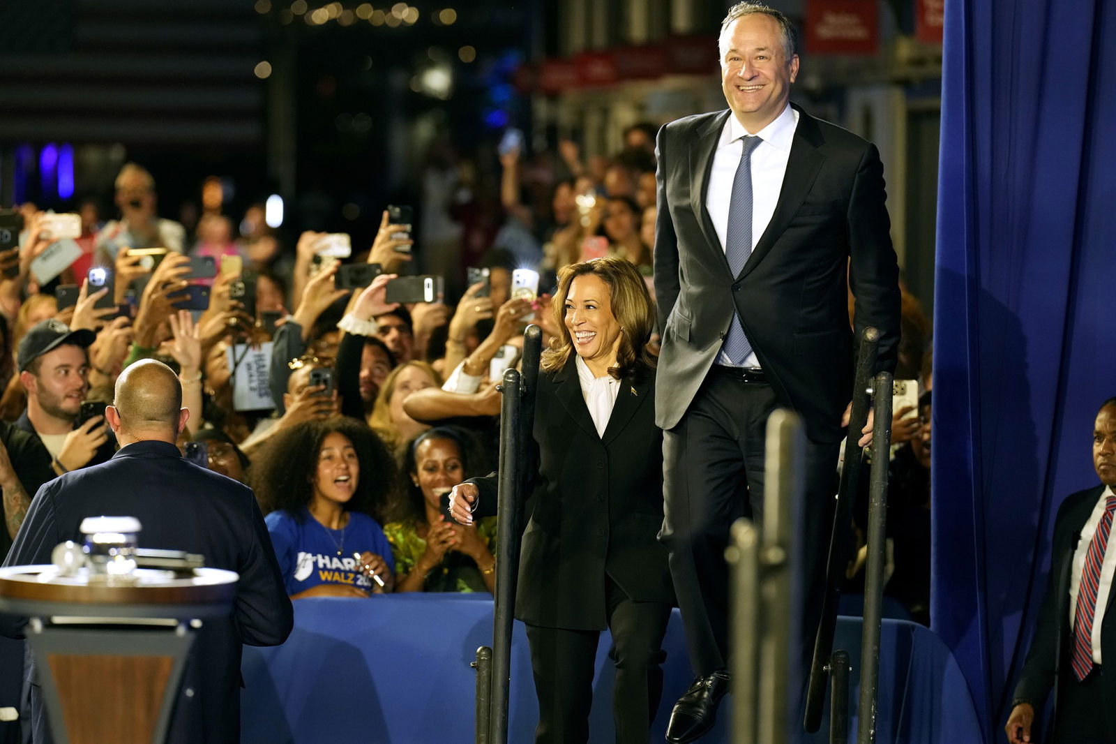 A man and a woman in suits smile as they step onto a stage in front of a cheering crowd.