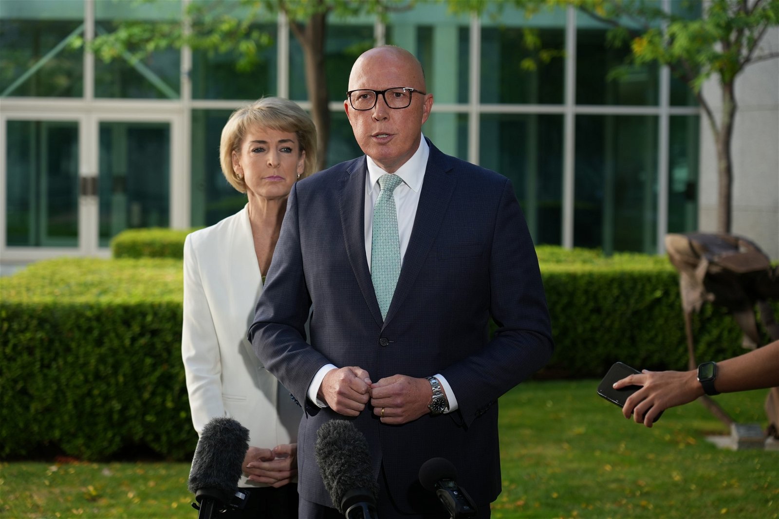 Peter Dutton speaking at a microphone with a woman standing behind him.