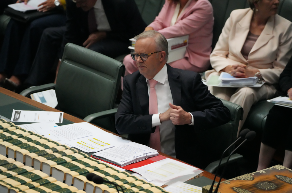 A man with grey hair, a pink tie and glasses sits down and reaches into his suit pocket.