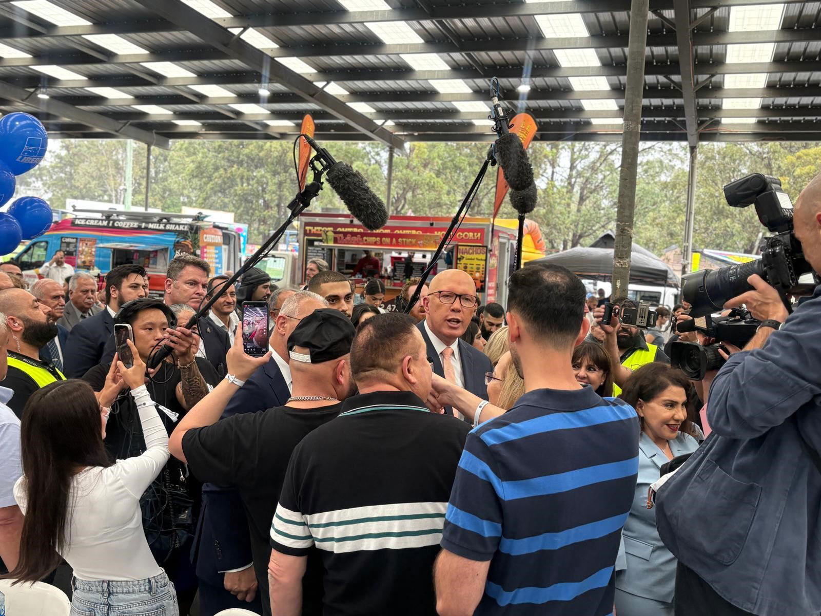 Peter Dutton is surrounded by a crowd, some carrying orange balloons and others boom microphones.
