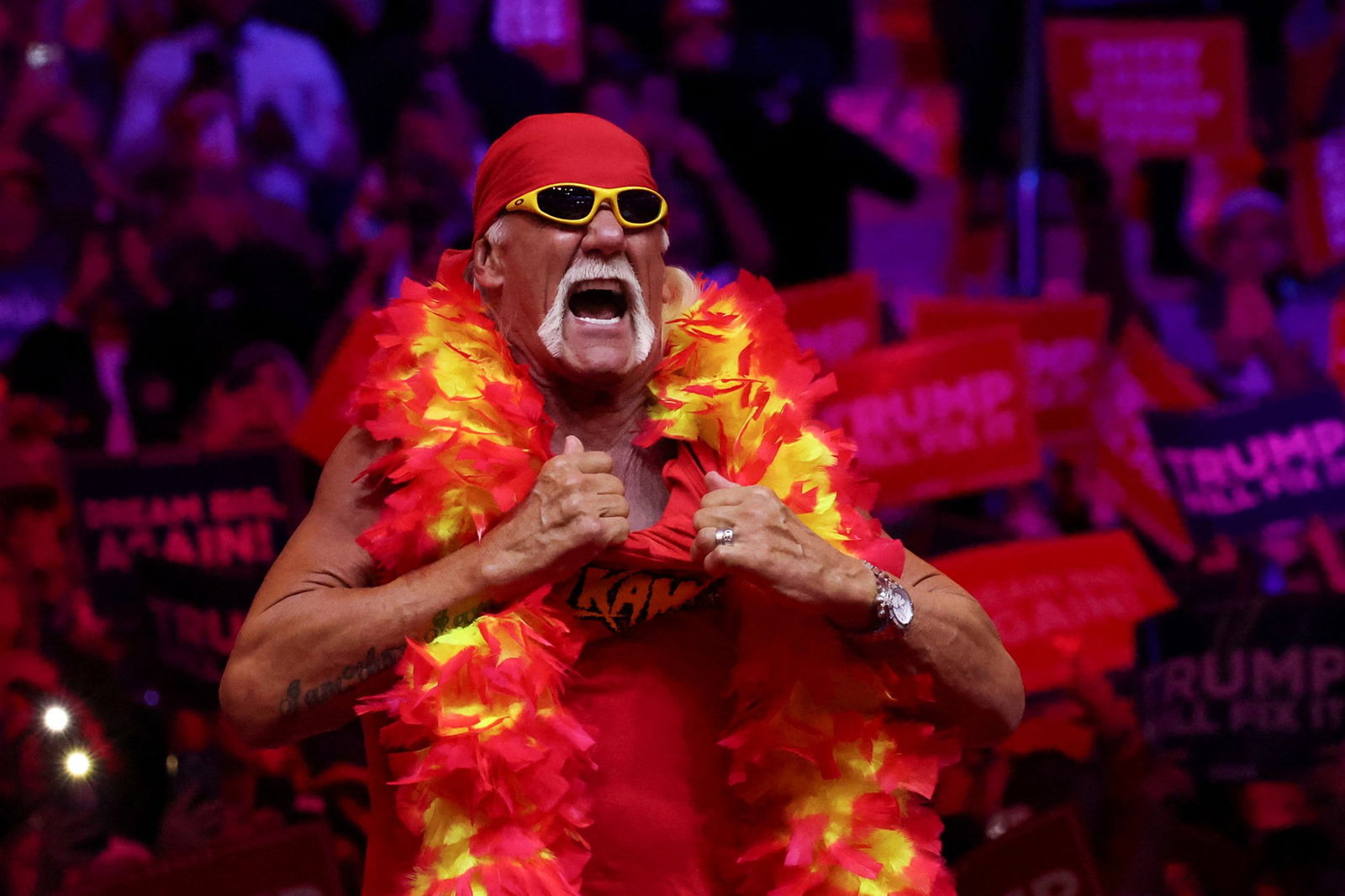 Hulk Hogan grabs at his singlet collar on stage. He wears a red and yellow feather boa