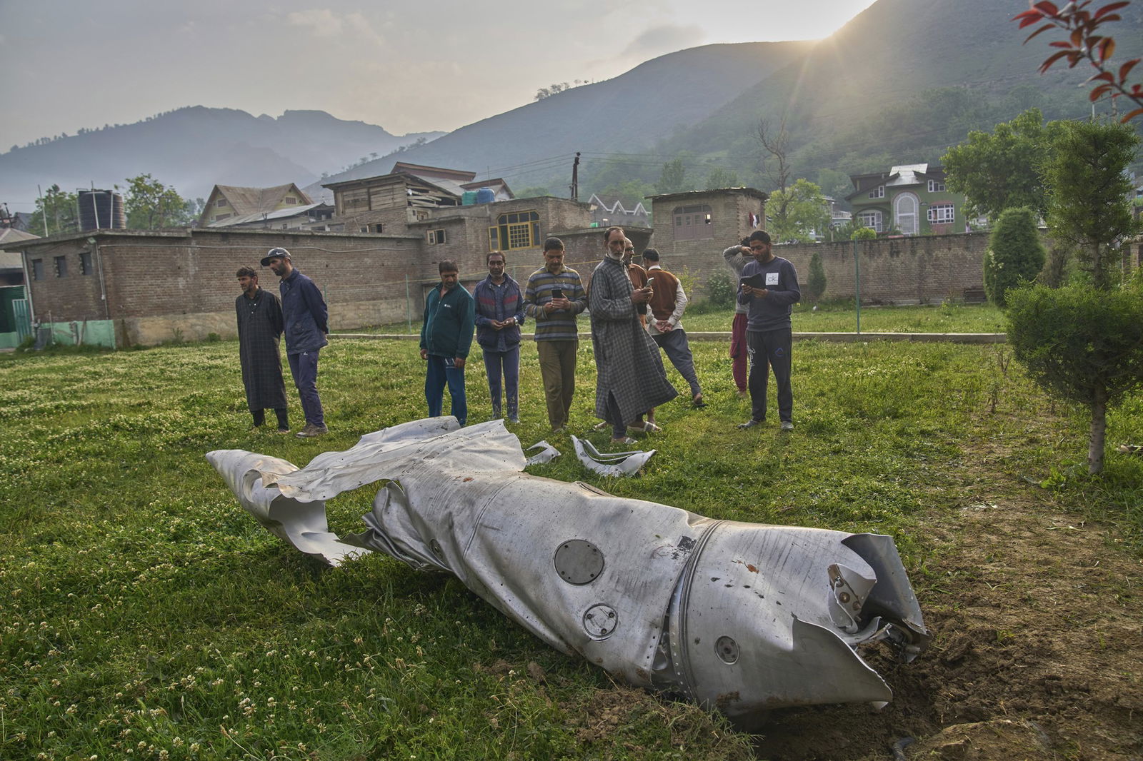 A damaged cylindrical piece of metal sits on grass with men gathered around looking at it