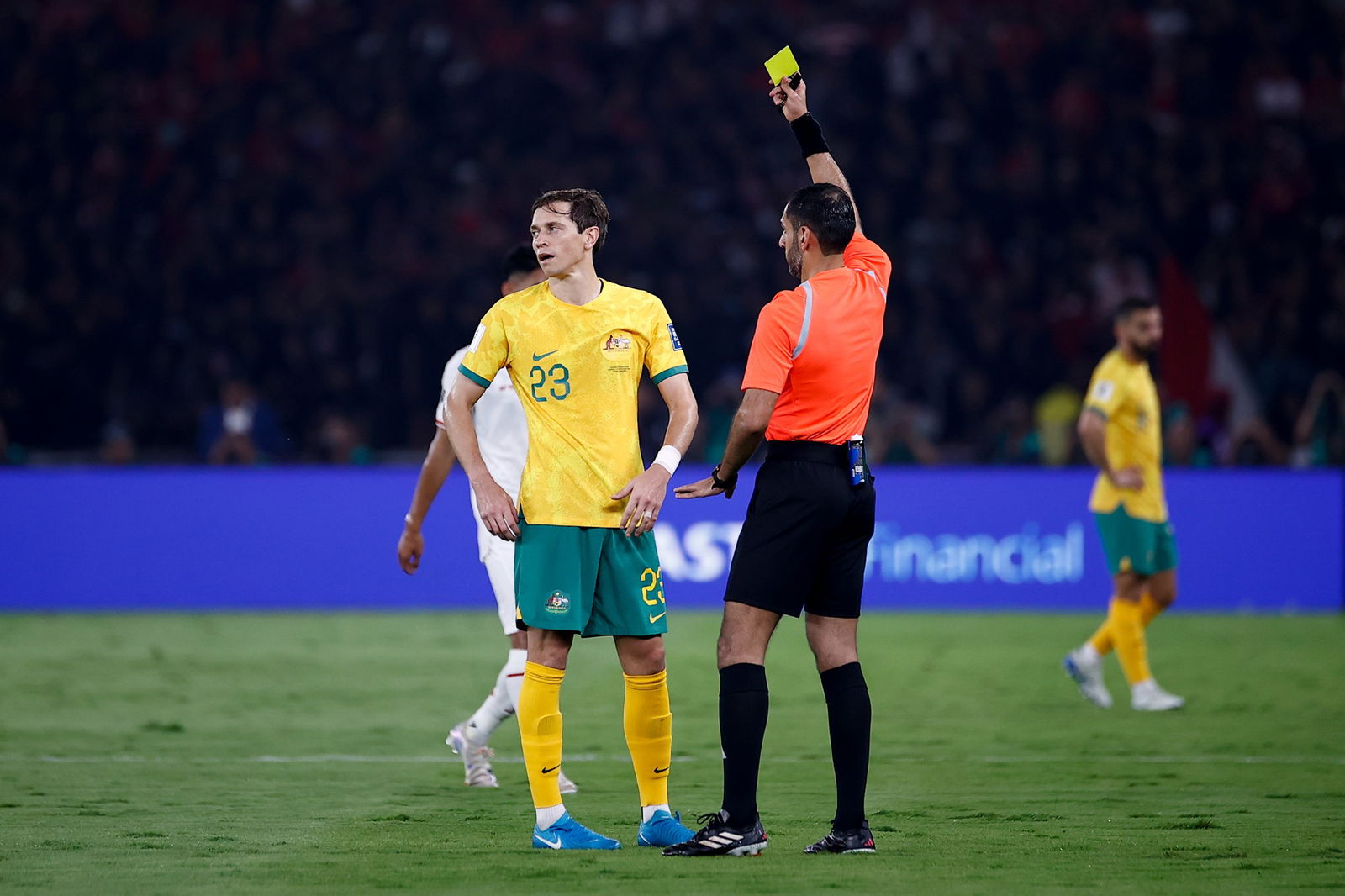 A soccer referee in an orange shirt holds up a yellow card to a player in green and gold, who looks away and has hands on hips.