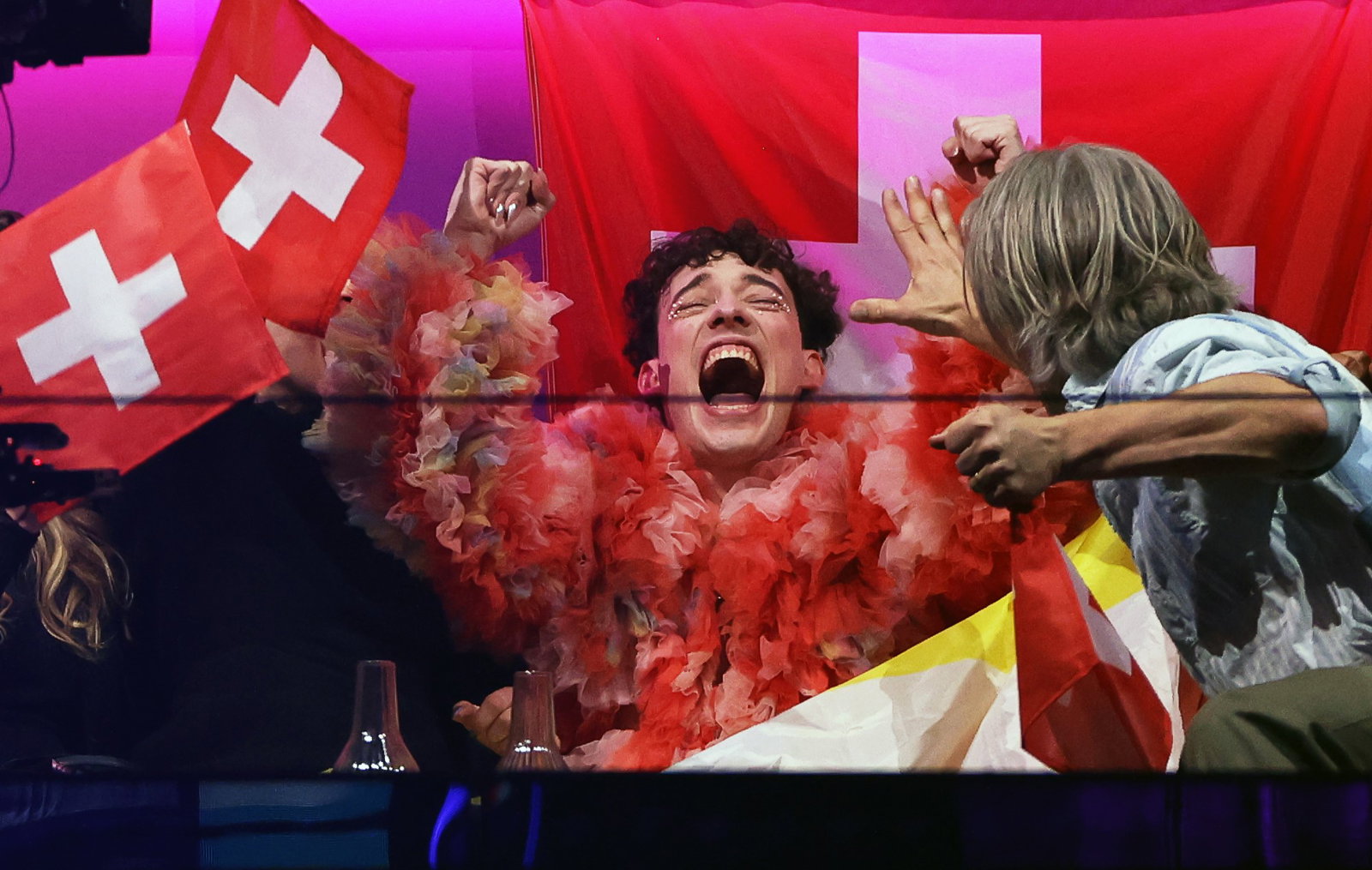 Artist nemo in red cheering as he wins Eurovision with the Swiss flag next to him