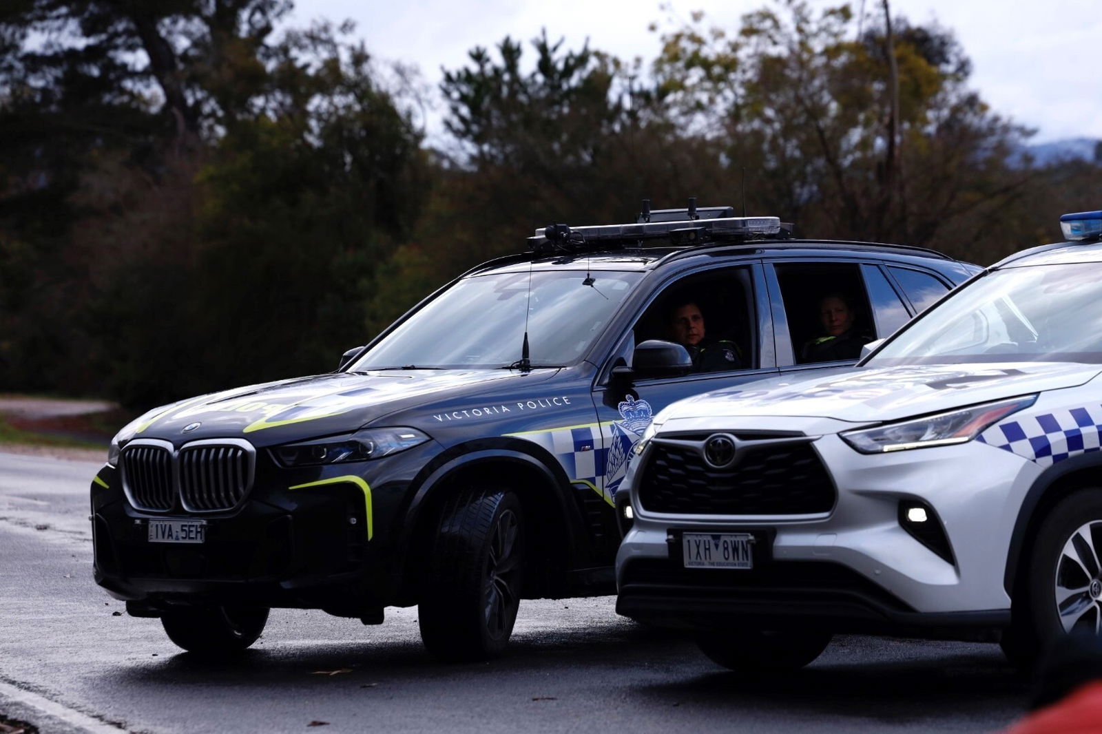 Two police officers look out of a dark police car that's parked beside a white and blue police car.