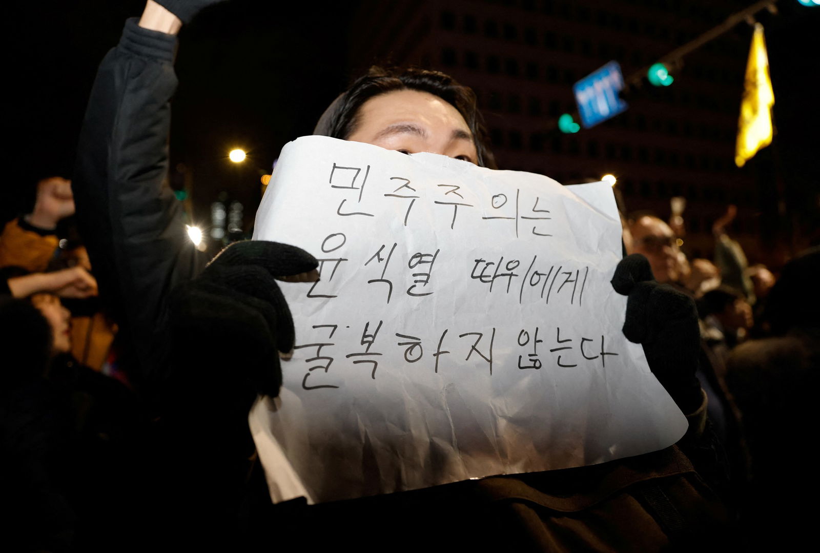 an Asian man in black in a crowd holds up a piece of paper with Korean characters on it