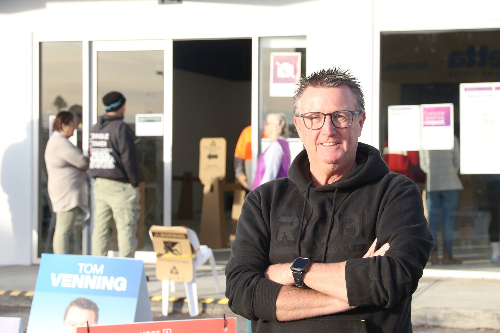 man in black hooded jumper stands with his arms crossed outside a polling location