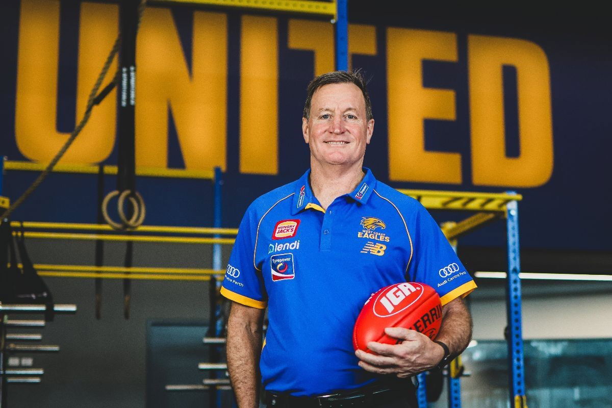 John Worsfold in a gym, holding an AFL ball, smiling