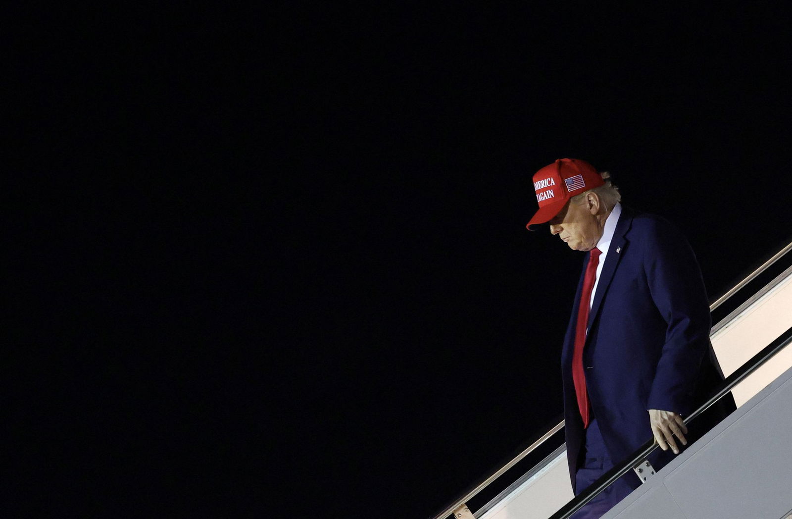 Donald Trump in a blue suit, red tie and red Make America Great Again hat descending an aircraft stairway at night with his left hand on a rail