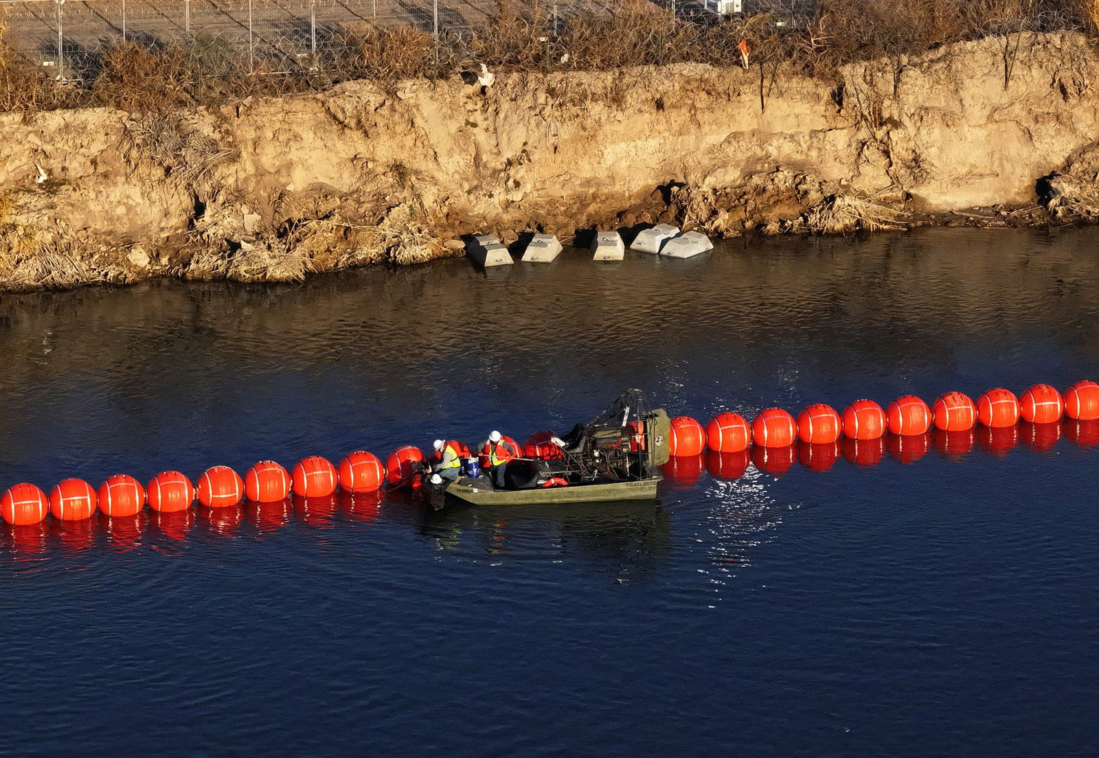 A small olive green boat alongside a row of red, circular buoys in dark river water alongside a rocky embankment