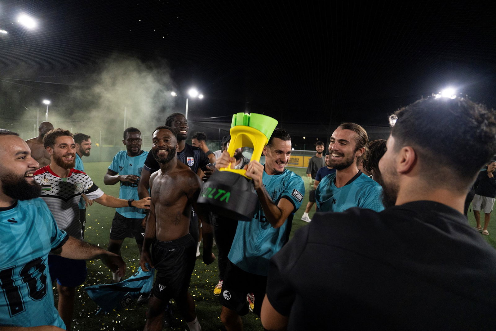 Um time de futebol comemora uma vitória formando um círculo ao redor de um troféu sorrindo.