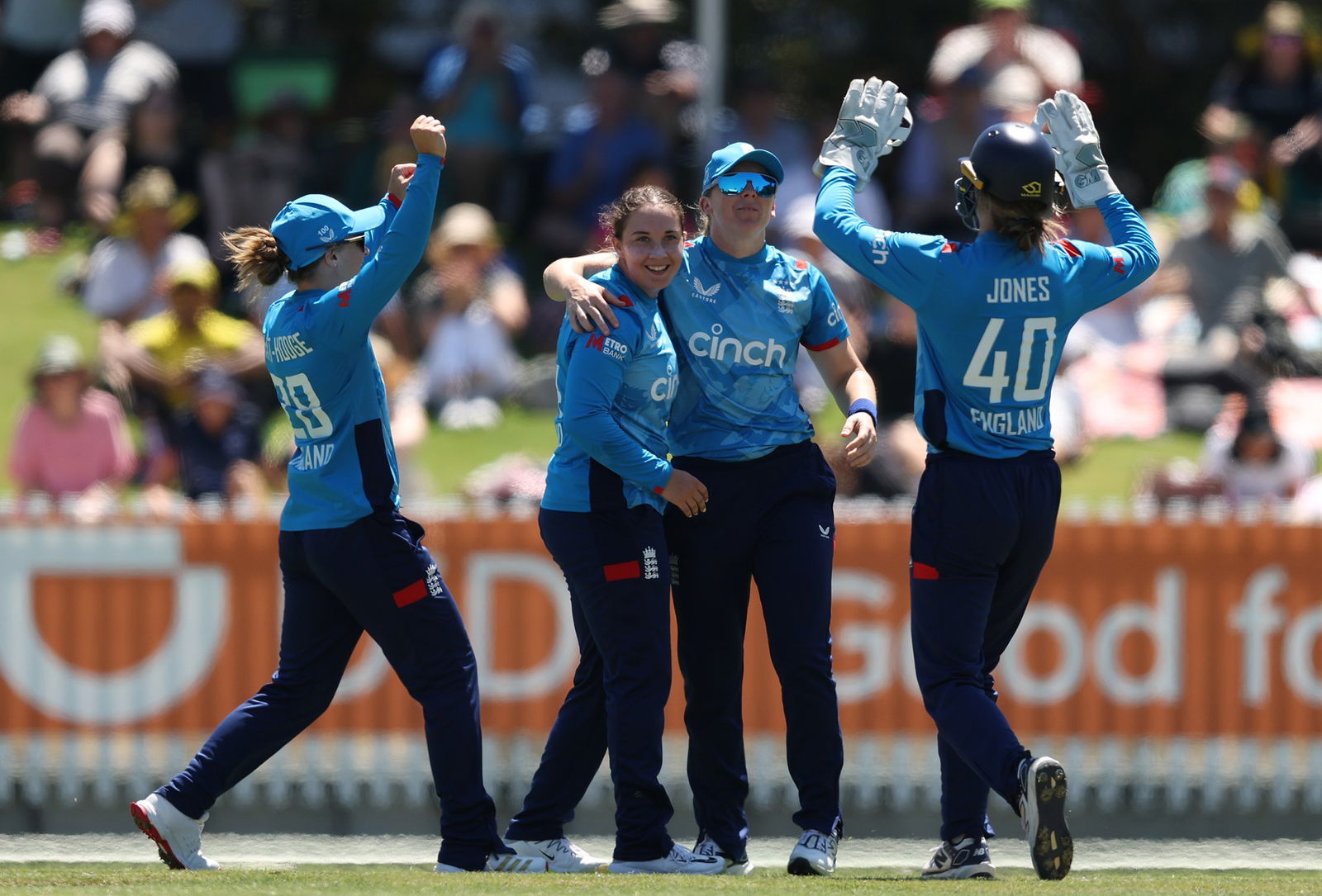 Four cricketers in blue shirts and long navy blue pants celebrate a wicket in bright sunshine in front of advertisement hordings and a crowd.