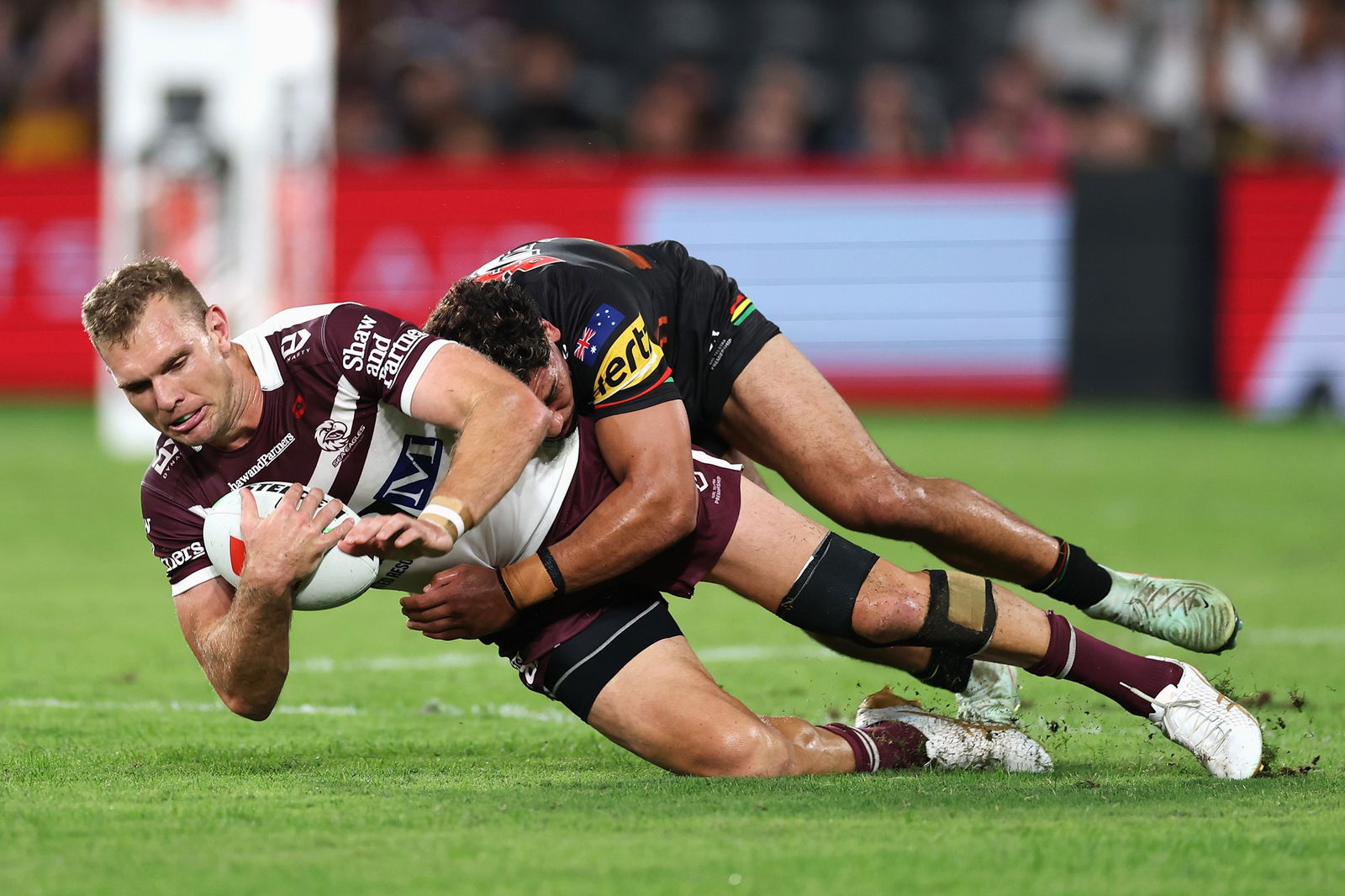 A rugby league player wearing maroon and white dives with the ball with a player in black tackling him from behind