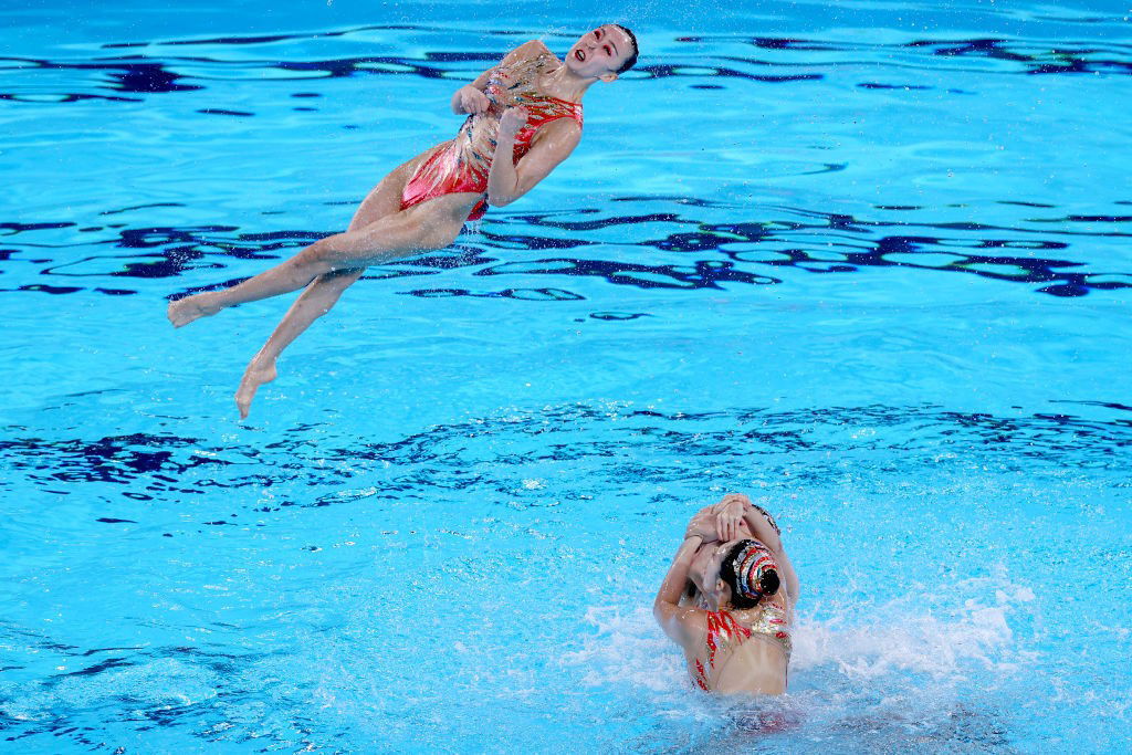 A woman pirouettes in mid air above two swimmers in a pool.