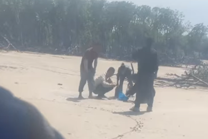 Four men gather around a bag of water bottles on a beach.
