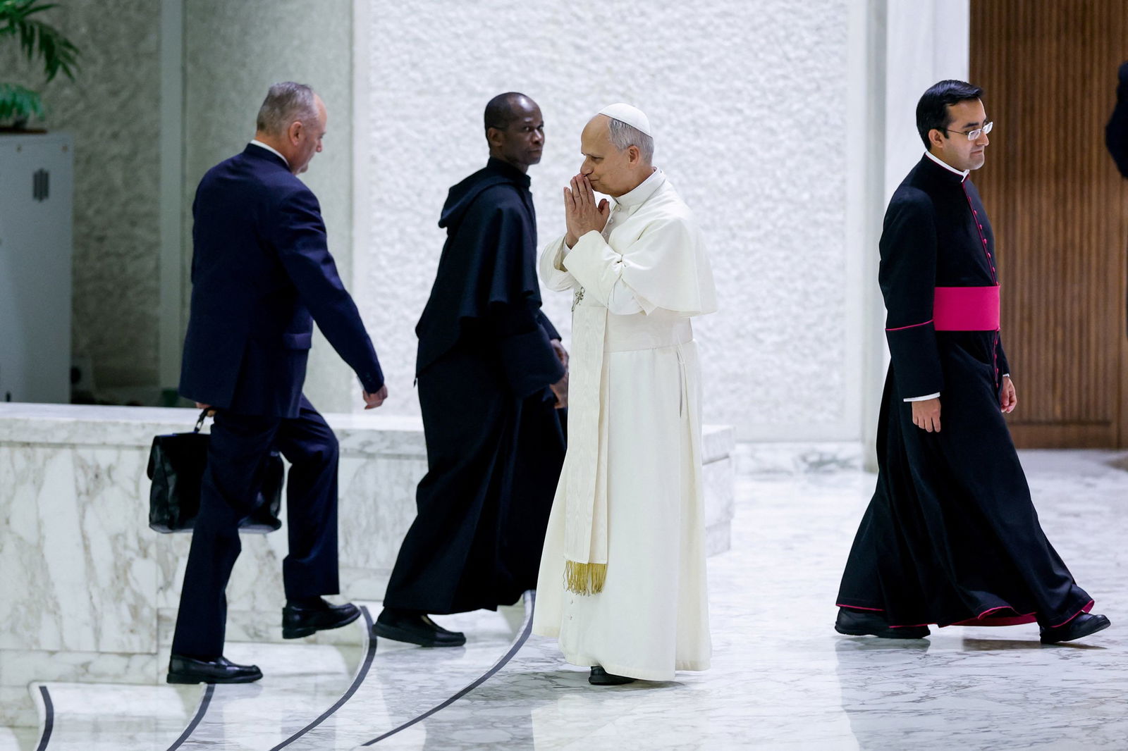 Um homem com um vestido branco majestoso e um chapéu junta as mãos em agradecimento na frente da boca enquanto olha para as pessoas fora de cena.
