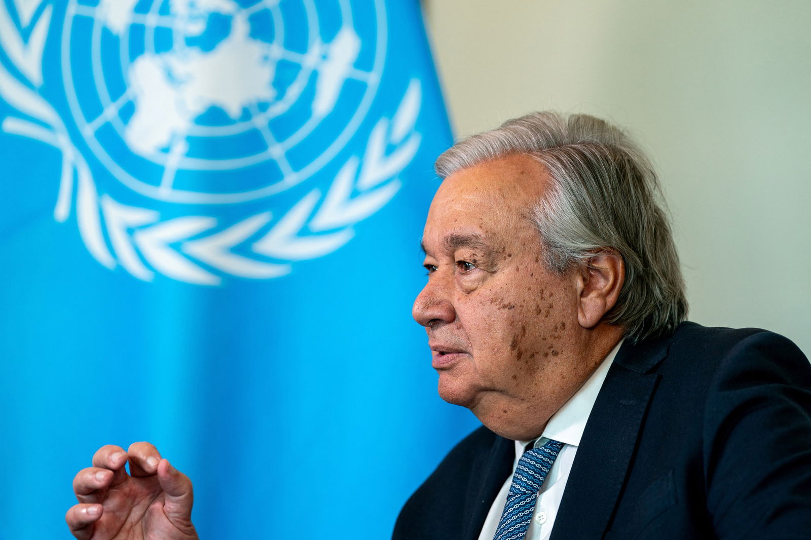 A close-up side profile of an older, grey-haired man in a suit inf front of the UN flag