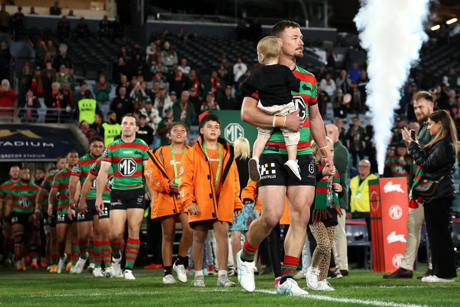 Damien Cook leads the South Sydney Rabbitohs onto the field for an NRL game while carrying his kids.