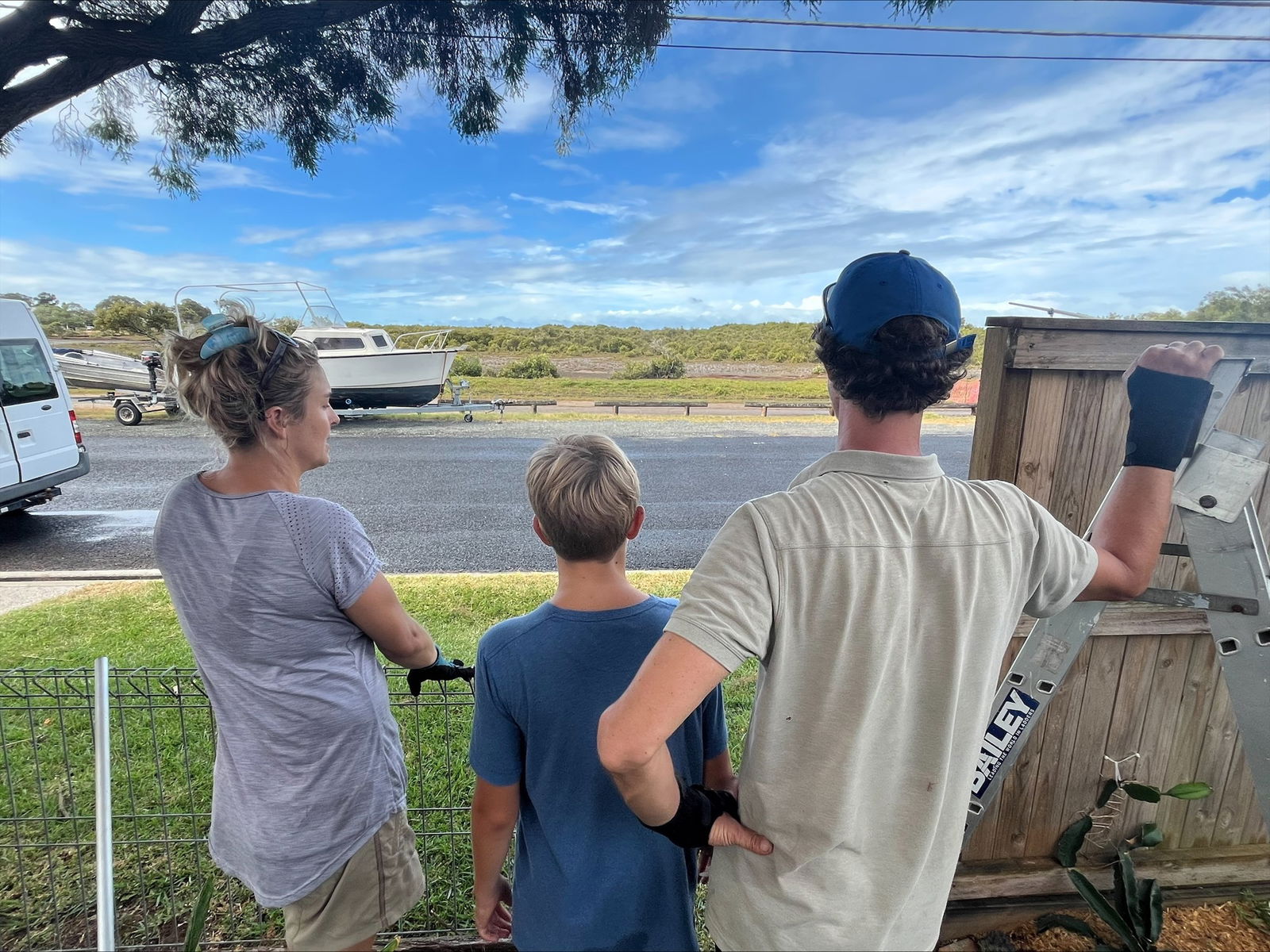 A man and a woman standing at their fence with their son looking out at mangroves and a blue sky.