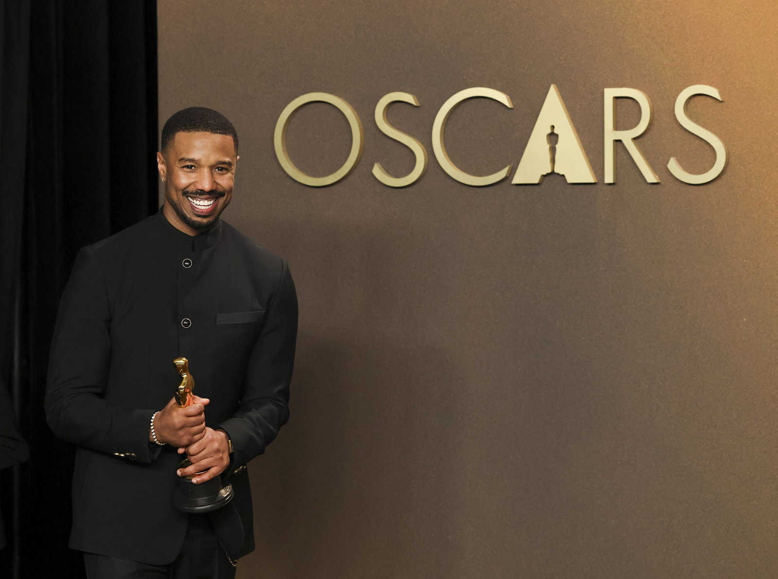 A man dressed in black, smiles as he holds his gold Oscar in both hands. The word Oscars is written on the wall behind him.