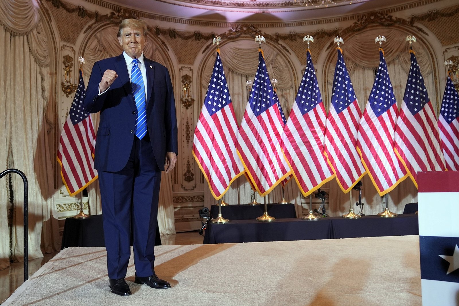 Donald Trump stands on a stage in front of a row of American flags. He is wearing a suit and holding up a fist.