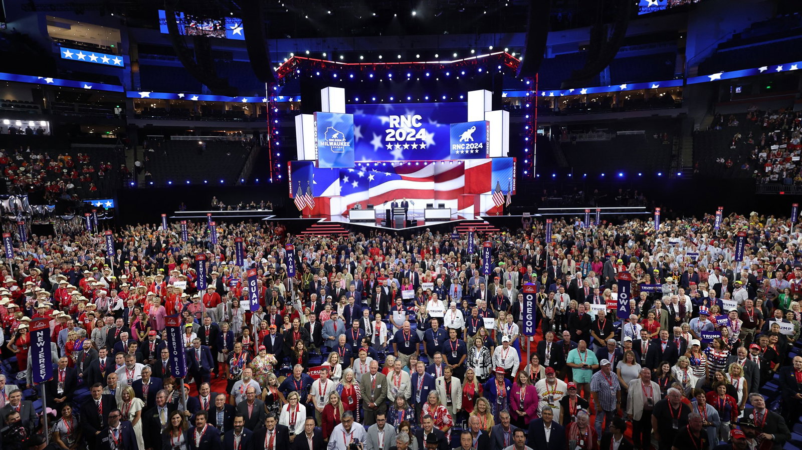 A crowd of people stand in front of a stage decorated with an American flag. 