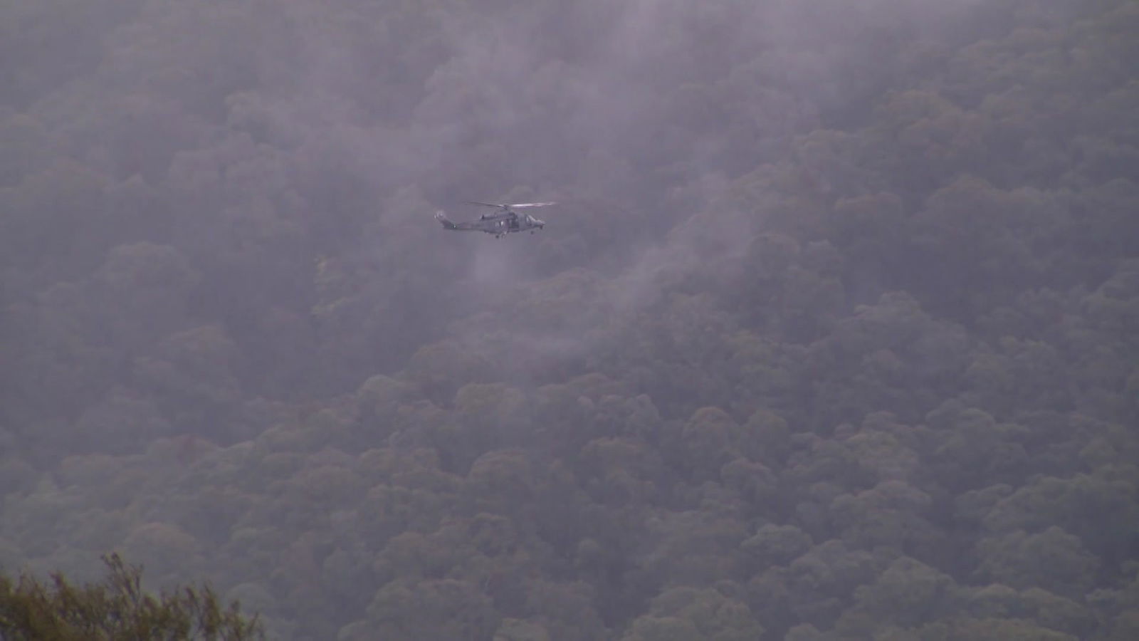 A dark coloured chopper flies through fog in front of a tree covered mountain.