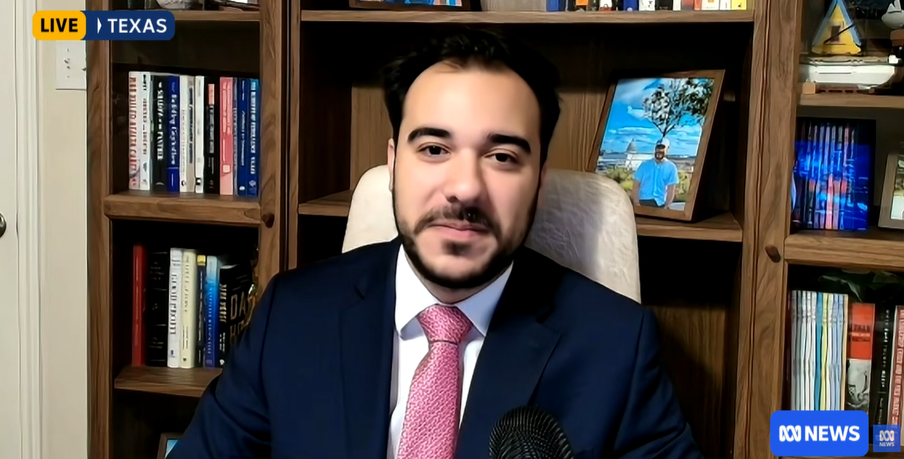 Um homem de terno azul, camisa branca e gravata rosa olha para a câmera enquanto está sentado em frente a uma estante 