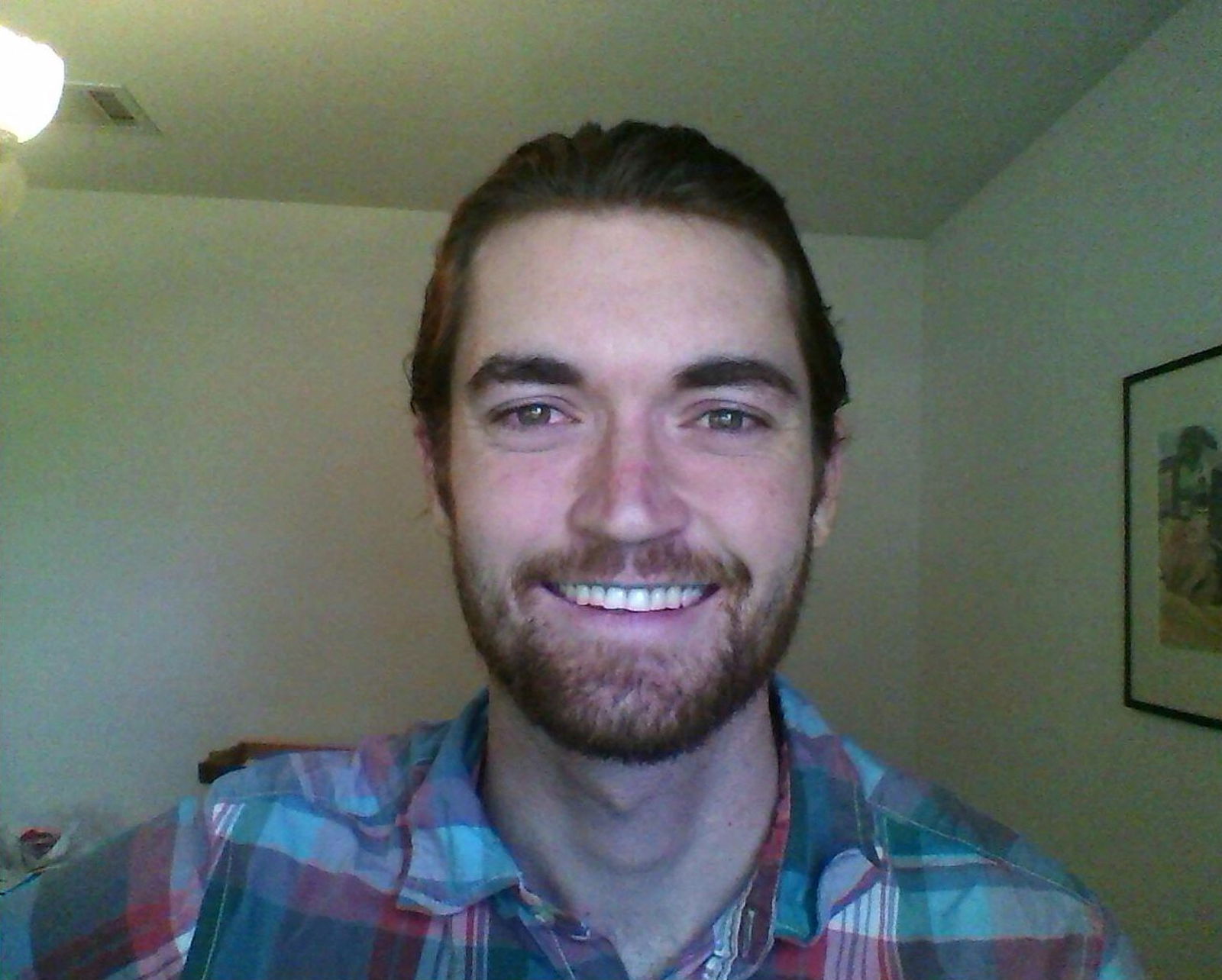 A man with brown hair and stubble, wearing a blue and red checked shirt while smiling