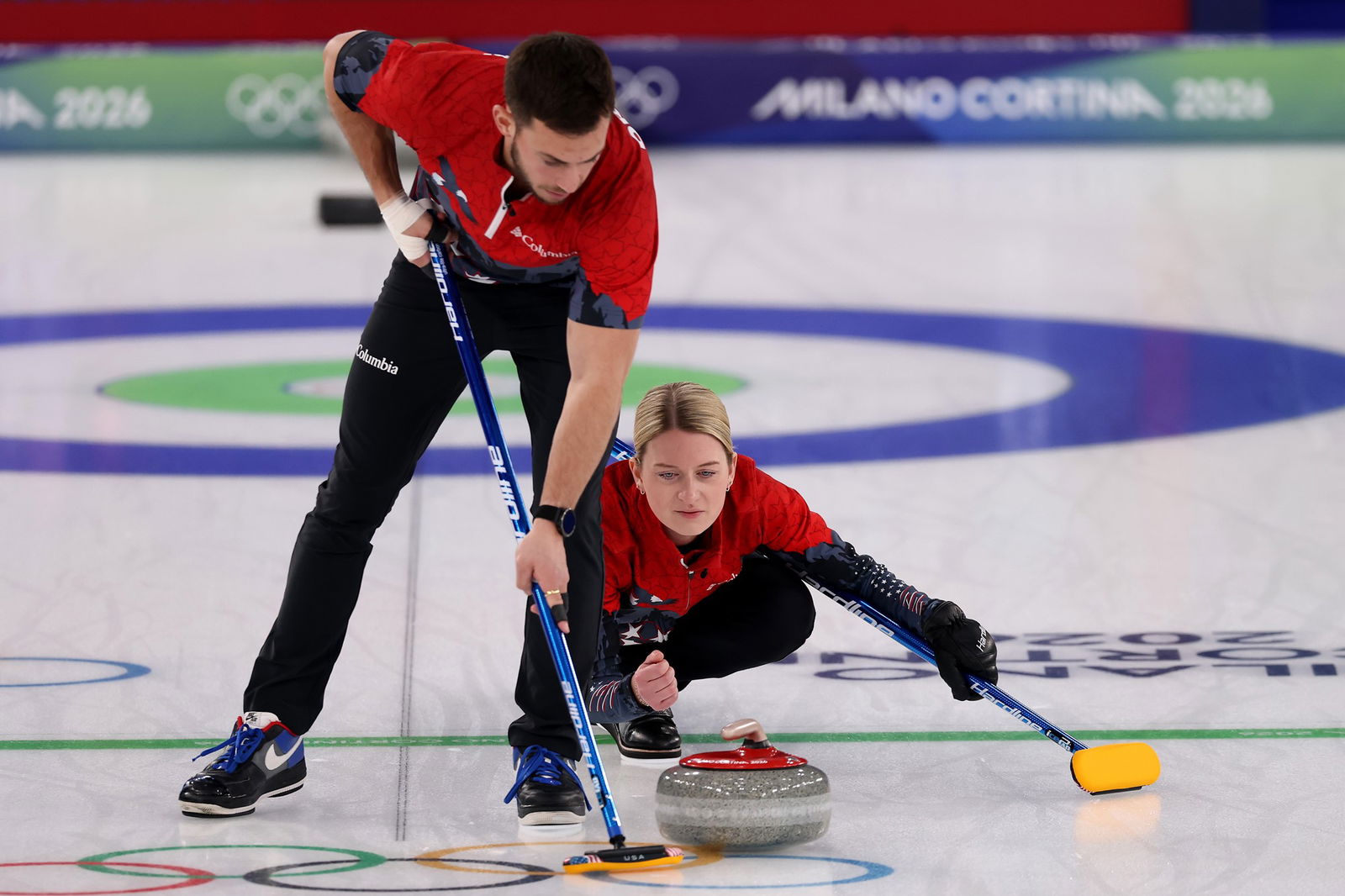 Two athletes in red and black are playing curling on an icy surface