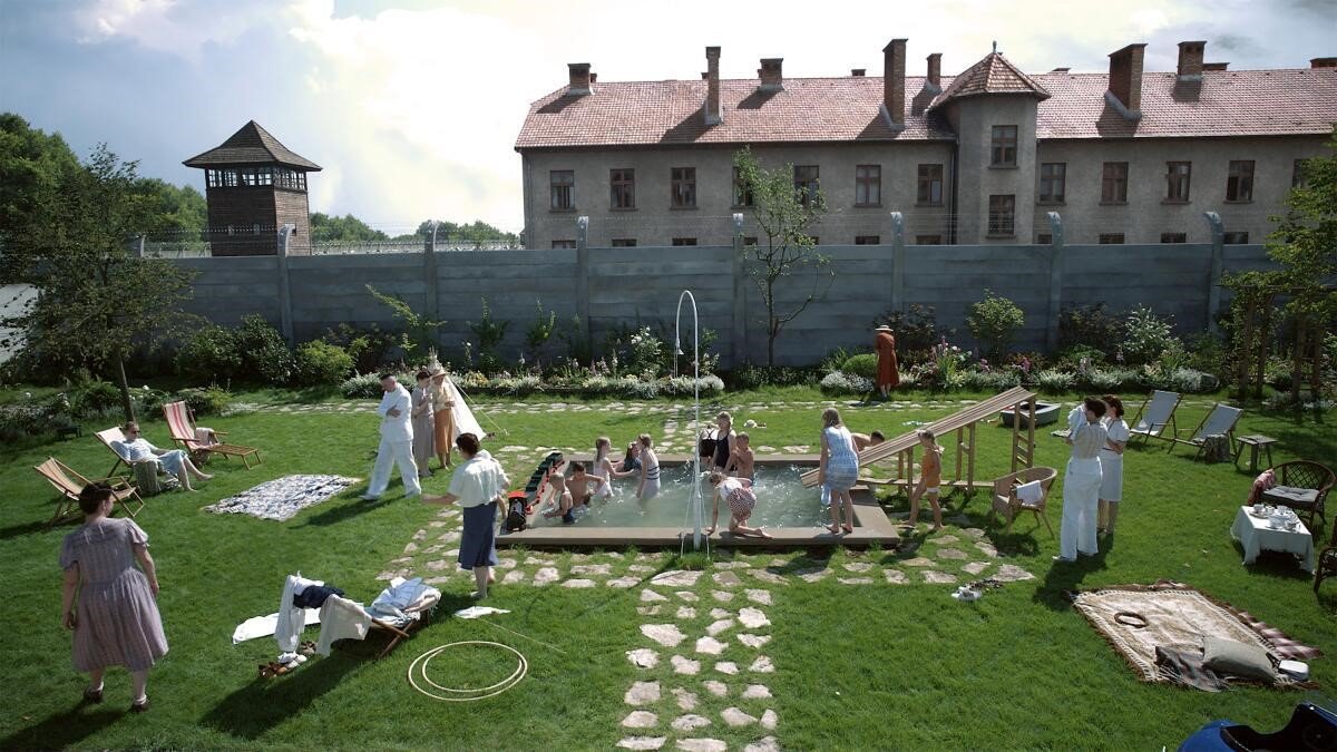 Still from The Zone of Interest, featuring a beautiful green garden and a pool filled with children, with Auschwitz in the background behind a wall.