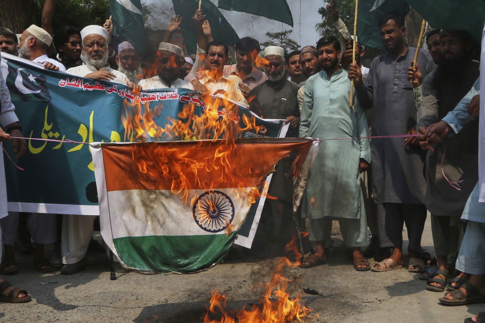 An Indian flag burns which being held by two people at a large protest with men gathered around watching it