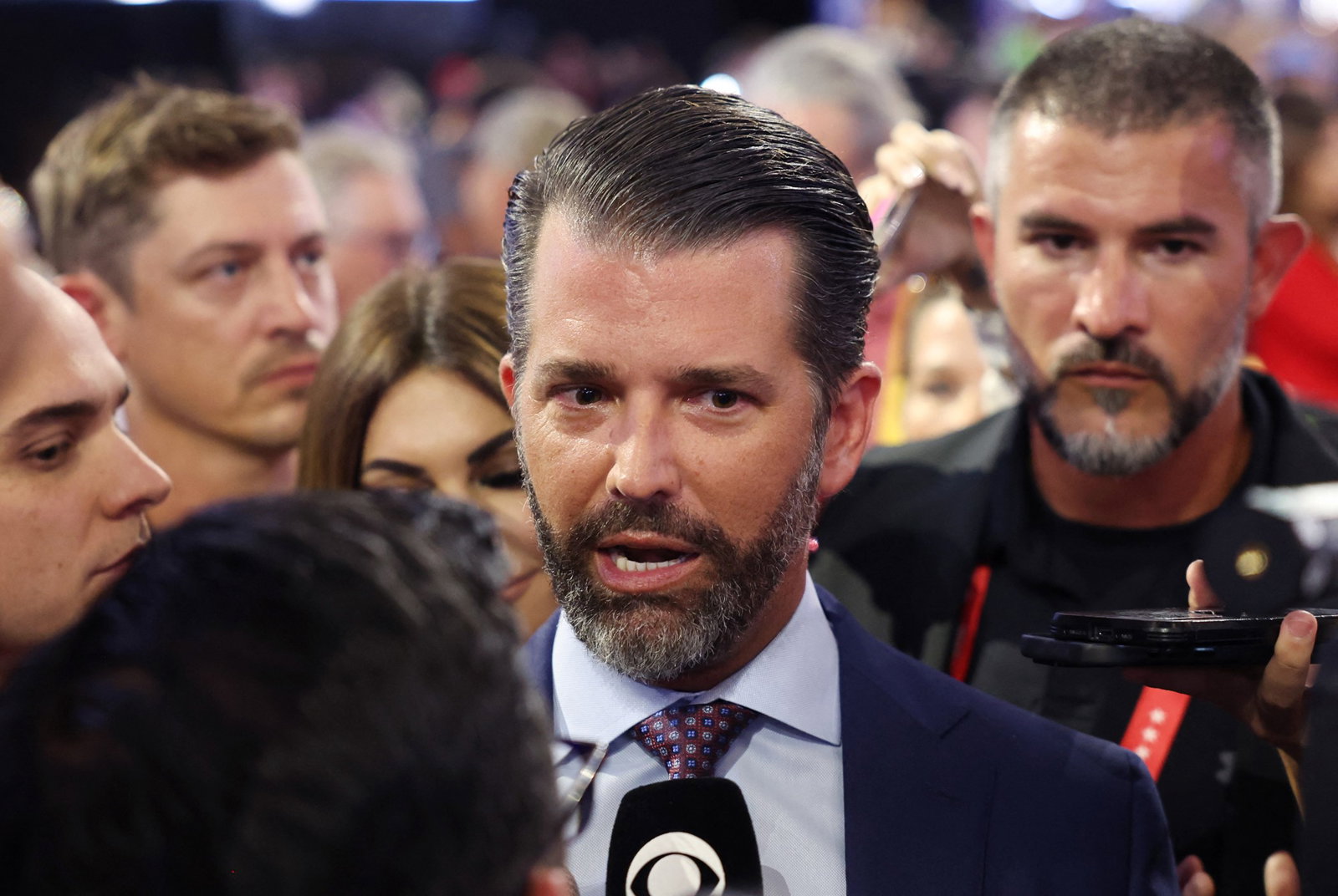 Donald Trump Jr speaks into a reporter's microphone. There's a crowd behind him.