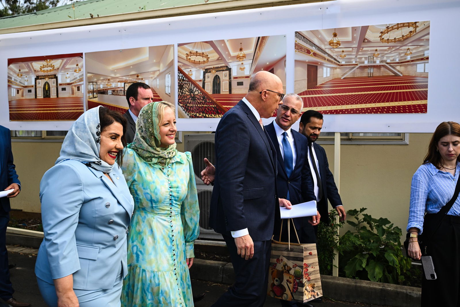 Peter Dutton walks in front of sign showing pictures of what appears to be the inside of a mosque.