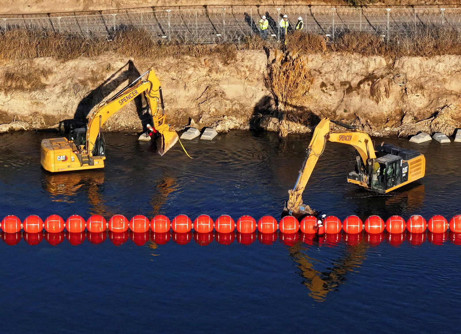 Two yellow machine diggers alongside a row of red, circular buoys in dark river water alongside a rocky embankment