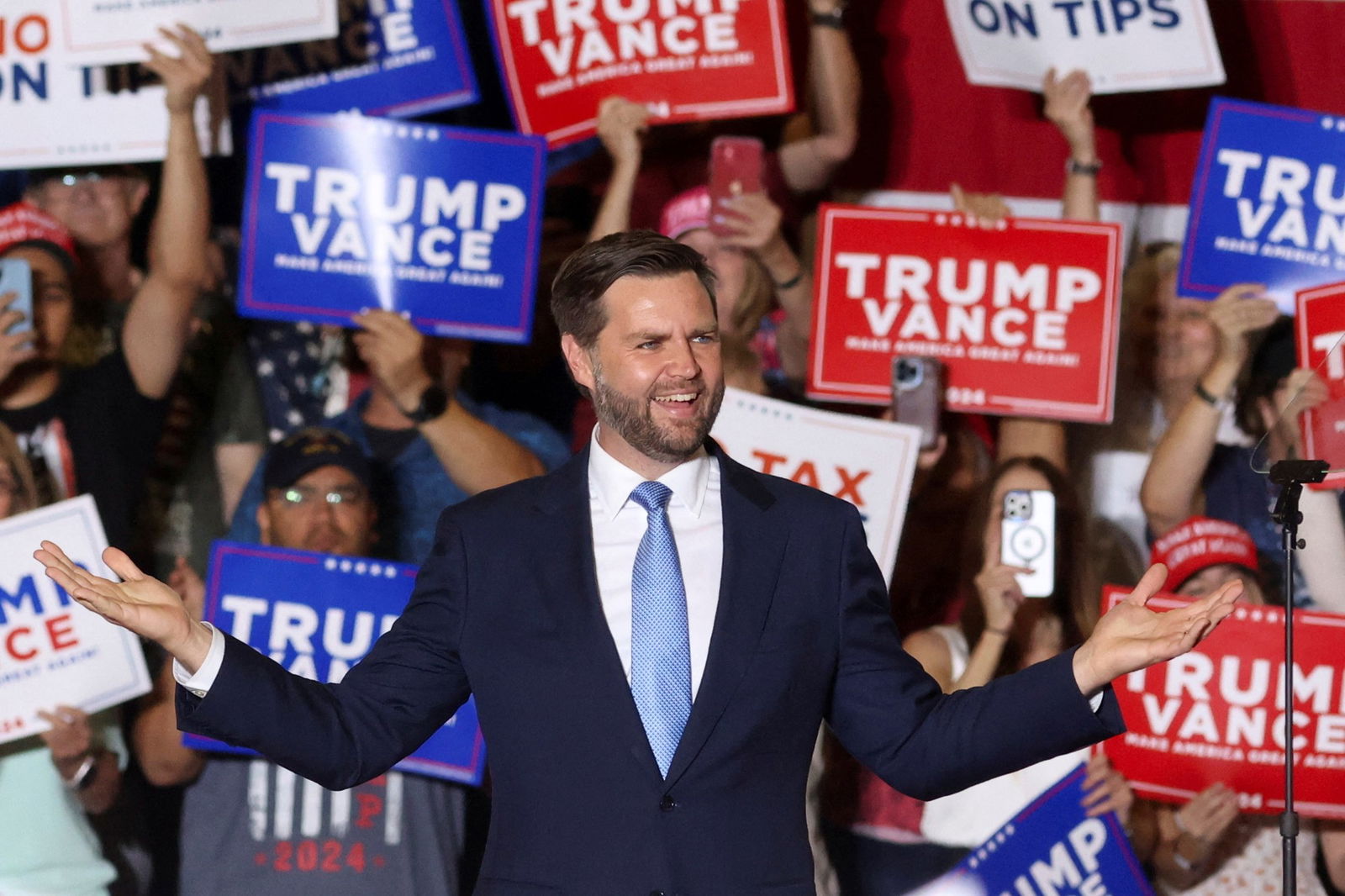 JD Vance stands in front of a crowd with “Trump Vance” signs.