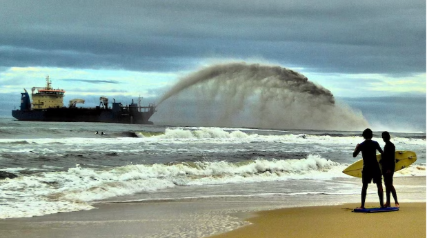 A large boat spraying huge amounts of sand towards the beach in a bug arc. Two surfers in the foreground. 