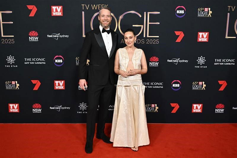 43 yaşındaki Hamish Blake, bir smokin ve bow-tie'de ve 45 yaşındaki Zoë Foster Blake, bej bir bağlantı üstünde ve eşleşen etekte, medya duvarının önündeki Logies kırmızı halısına gülümseyen poz veriyor.