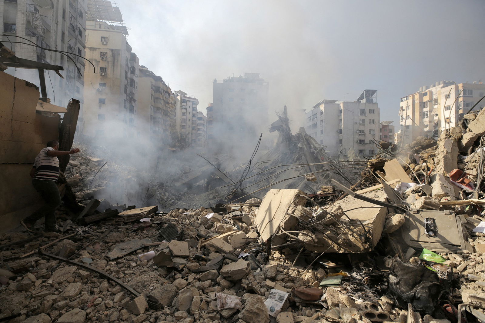 Smoke rises from rubble in the foreground, buildings in the background