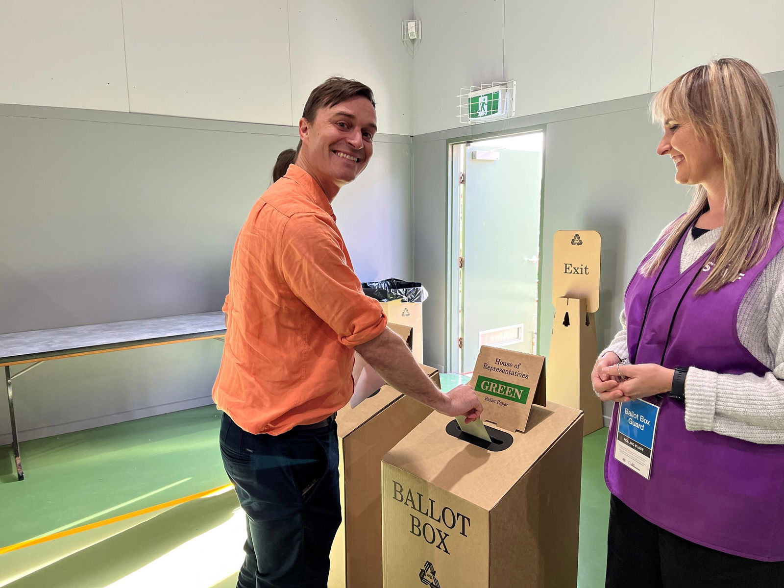 Alex Dyson wearing an orange linen shirt and smiling while placing a voting ballot into a carboard ballot box