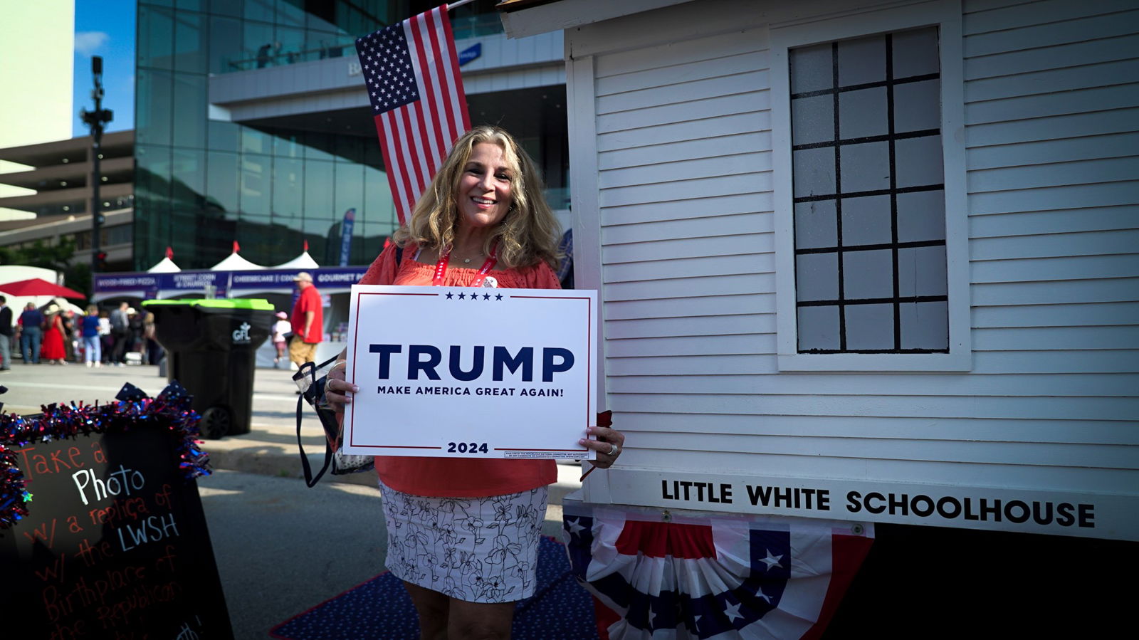 A woman wearing a white skirt and red top holds a TRUMP sign standing in front of a US flag