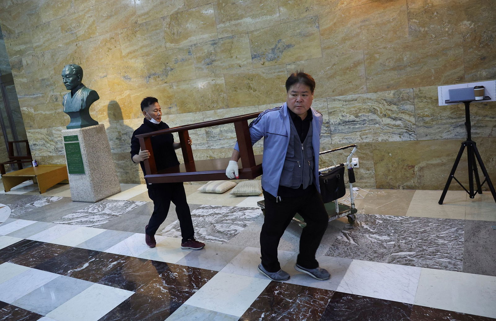 two Asian men carry a bookshelf in a marble foyer looking dejected