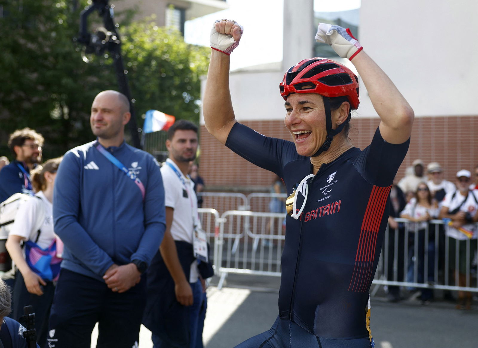 A woman wearing dark blue Lycra with a red cycling helmet has her arms raised in celebration. She is smiling. There is a crowd behind her, she is seperated from them by a portable fence.