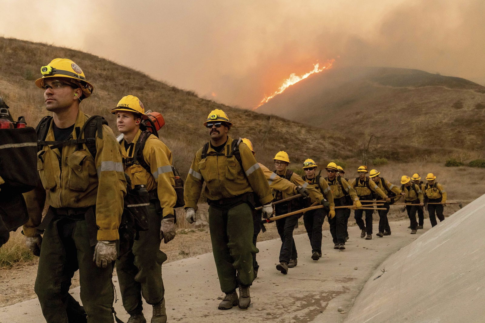 A line of firefighters on a dirt track, with a burning hill behind them