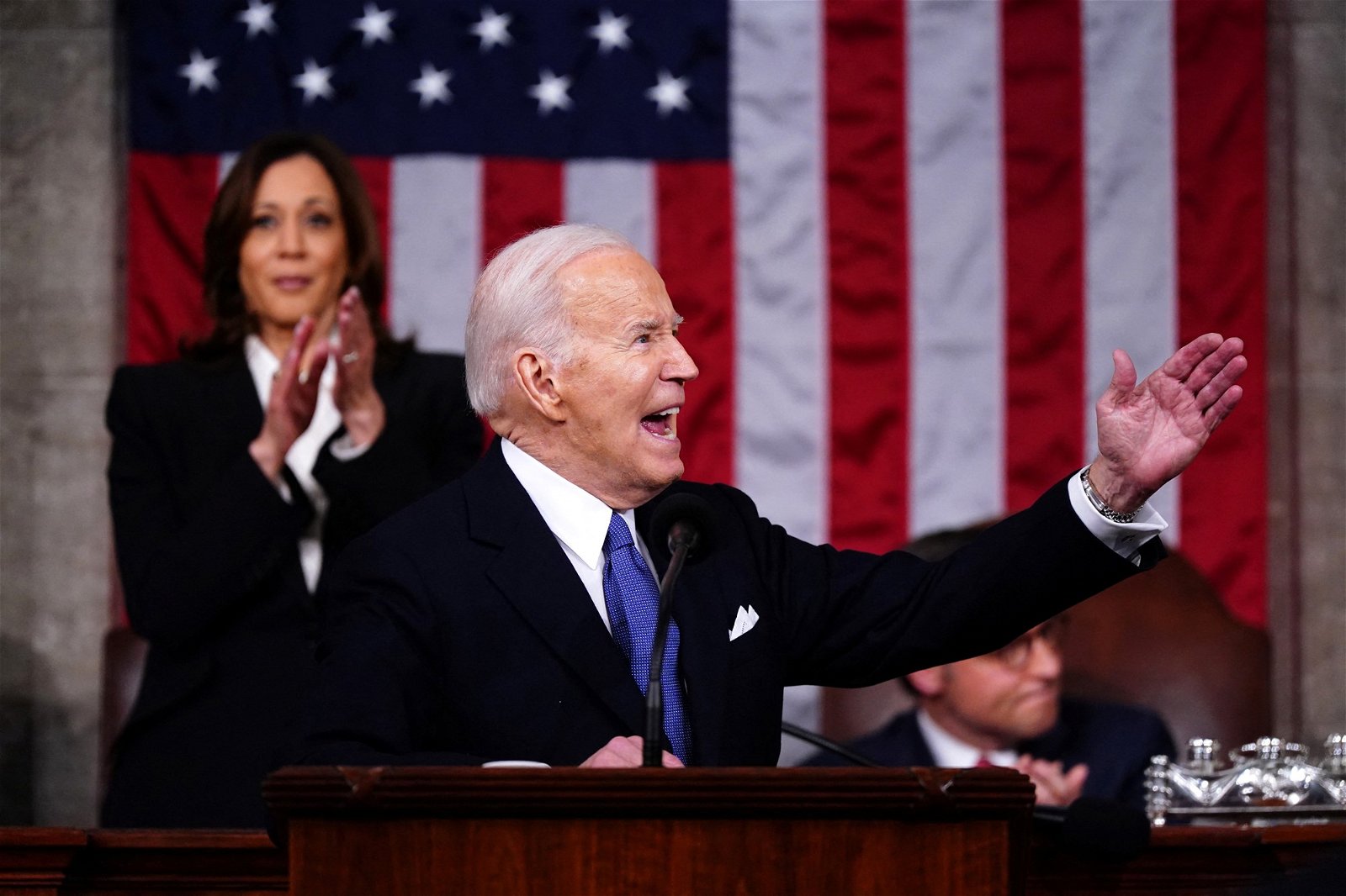 Joe Biden holds one hand out as Kamala Harris claps behind him, a US flag on the wall