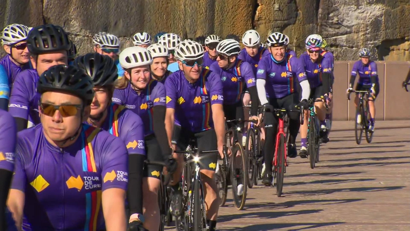 Decenas de ciclistas con lycra violeta y cascos de bicicleta viajan.