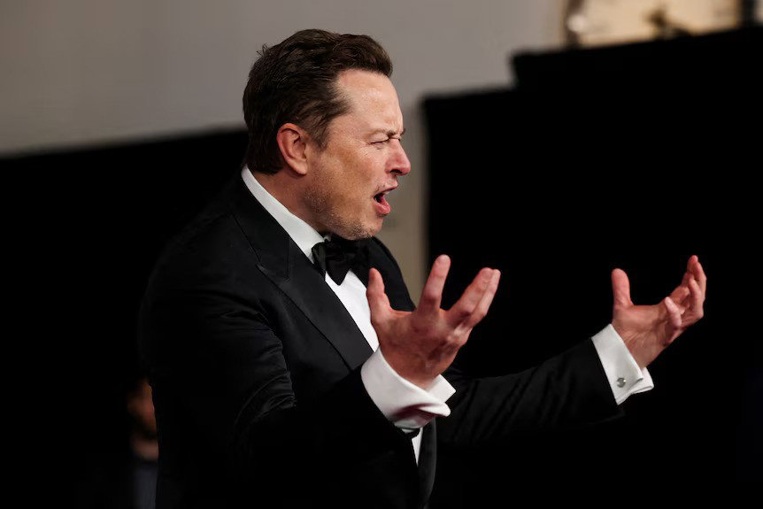Musk in a black tie suit scrunching his face and hands