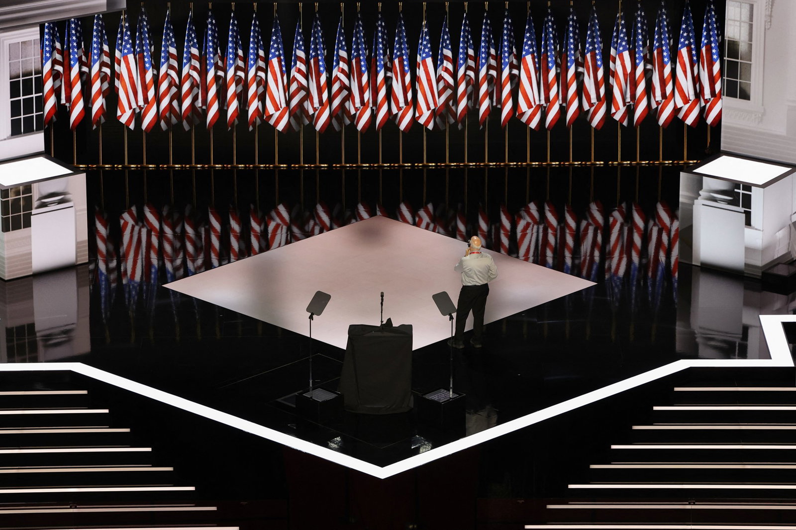 A man stands on an empty stage in front of a long row of American flags.